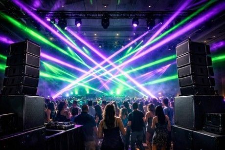 Concert crowd under crossed purple and green laser lights, with large speakers on both sides.