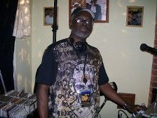 Man with glasses and headphones at a DJ setup, wearing a skull-print shirt.