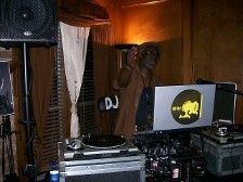 DJ at a venue, mixing music. DJ wears glasses, brown outfit, hand raised. Equipment lit, dark setting.
