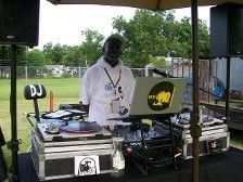 DJ stands behind his equipment, set up outdoors under a tent, ready to play music.