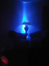 Blue-lit lamp on a table in a dark room, beside a shadowy armchair.