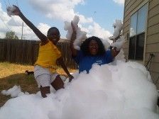 Two Black women laughing, playing in a large pile of white foam outdoors. One wears yellow, the other blue.