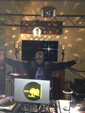 DJ with arms outstretched in front of a console with records, inside a room with decorative lights.