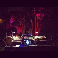 DJ setup with turntables, laptop, and lights, in a dimly lit venue with a pink hue.
