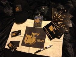 Black merchandise with a gold logo: hat, patches, cards, lanyard, pen, on a tan cloth.