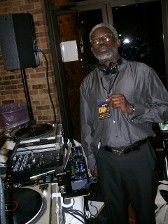 Man with gray hair DJs in a dim room, wearing a gray shirt and headphones.