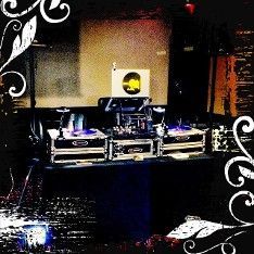 DJ setup in a dark club: turntables, mixer, and speakers on a black table with a white sign.