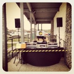 DJ setup outdoors: speakers, console on black table, yellow caution tape, under a covered area.