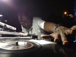 DJ in white shirt, scratching a vinyl record, arm extended, dimly lit outdoor setting.