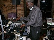 DJ, older Black man in grey shirt, mixing music at a dark event, with headphones around his neck.