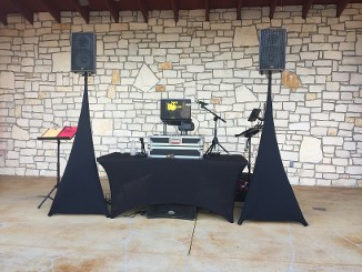 DJ setup with speakers, equipment on a black-covered table, and a microphone stand in front of a stone wall.