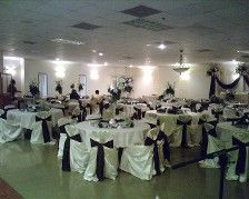Large banquet hall with round tables draped in white linens and black chair sashes, set for an event.