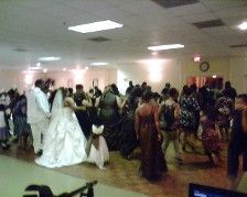 Wedding reception: Bride & groom dance, guests around. Wooden floor, white room with lights.