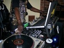 DJ at work: black man at turntable, laptop, mixing console. Indoor setting, dark clothing.
