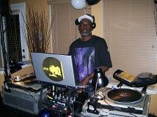 DJ, older Black man at a party with turntables, laptop and headphones, in a room with blinds.