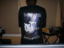 Person wearing a black shirt with a logo on the back, standing in front of a keyboard and speakers.