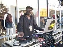 DJ, man in black, at outdoor event, turntable, laptop, and speakers, store window in background.