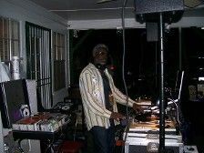 DJ wearing a striped shirt and glasses, stands at a DJ setup under a covered outdoor area.