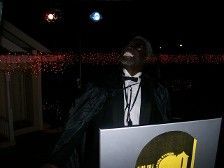 Man in tuxedo, looking up, at a DJ booth with a logo.