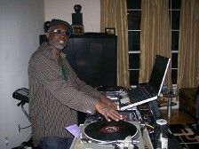 DJ spinning records indoors, wearing a hat and jacket. Laptop and window in the background.