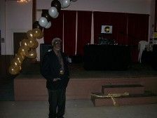 Man in a blazer stands on a stage decorated with balloons. There is a DJ setup behind him.