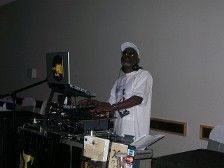 DJ stands at a music console, wearing a white cap and tee. Interior shot.