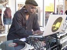 DJ, black man in black shirt and cap, working at laptop and turntable outdoors.