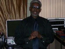 Man with gray hair and glasses in a black shirt and tie, hands clasped, smiling, in front of a computer.