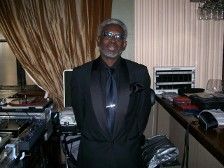 Man in a black suit, silver hair, and glasses, standing behind a DJ setup at a bar.