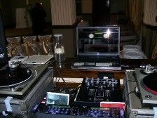DJ setup with turntables, mixer, laptop, and water bottle. Low light setting.