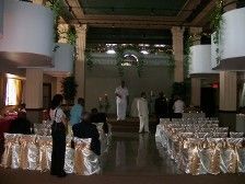 Wedding ceremony, indoor venue, people seated, officiant standing, decorated chairs.