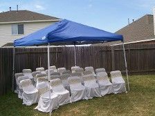 A blue tent set up over white chairs on a lawn.  A backyard setting with a wooden fence.