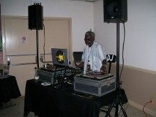 DJ at a party, mixing music on turntables with speakers. Elderly man in white shirt. Interior setting.