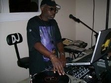 DJ in a cap, sunglasses, mixing on turntables with a laptop and microphone.