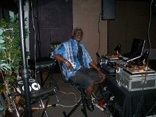 Man seated at DJ equipment, wearing blue shirt, shorts, and dark shoes, set up outside.