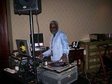 DJ at an event, man mixing music at a turntable, speakers and equipment, wearing a blue shirt, indoors.