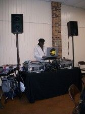 DJ at a party with equipment, laptop, and speakers. Man in white jacket, black table, brick pillar.