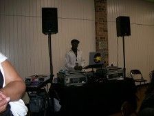 DJ at event, wearing a white shirt and cap, with speakers, turntables, and laptop set up.