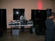 DJ at a party with equipment on a table, one person looking on, and a red light.