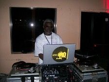 DJ at work, man behind turntables and laptop, in room with window. Laptop has yellow logo.