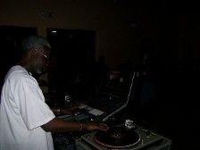 DJ at a dimly lit event, wearing white t-shirt, mixing on turntables with laptop, dark background.