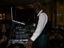 A DJ, an older Black man in a vest and jeans, standing at his equipment, likely at a wedding.