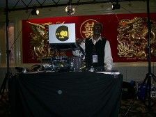 DJ standing behind his equipment at a red-themed event. He wears a black vest and a white shirt.