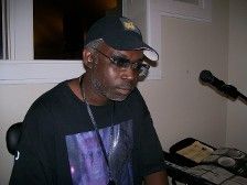 Man with glasses and a baseball cap sits at a keyboard, looking down. A microphone is visible.