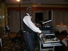 Man DJing at an event, wearing a black vest and jeans, operating equipment in a ballroom.