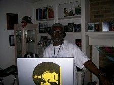 Man at DJ setup, wearing headphones and white shirt. Inside room, bookshelves in background.