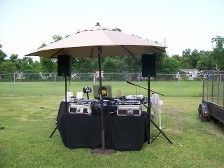 DJ setup under a beige umbrella on a grassy field, speakers, and equipment.