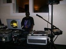 DJ at a party, seated behind equipment with a laptop, microphone, and turntables.