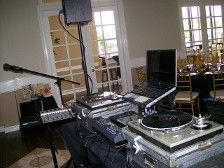DJ setup with turntables, mixer, microphone, laptop, and speaker in a room with windows.