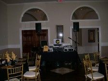 DJ setup in a room with speakers and tables. Gold chairs around tables with decor; neutral walls.